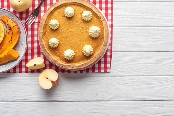 Delicious pumpkin pie with whipped cream on checkered napkin near sliced baked pumpkin, cut and whole apples, and fork on white wooden table — Stock Photo