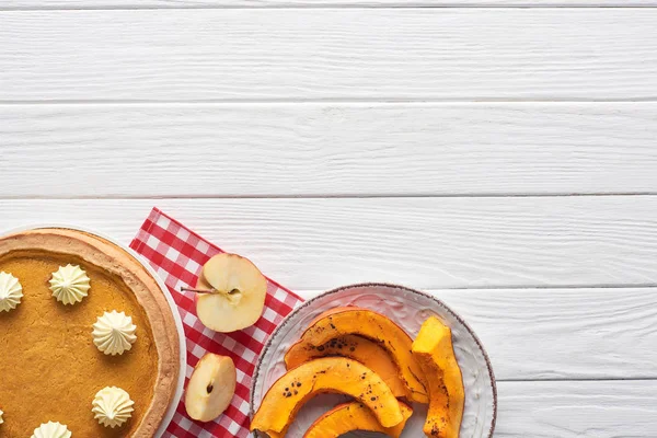 Délicieuse tarte à la citrouille avec crème fouettée sur une serviette à carreaux près de tranches de citrouille cuite au four et pomme coupée sur une table en bois blanc — Photo de stock