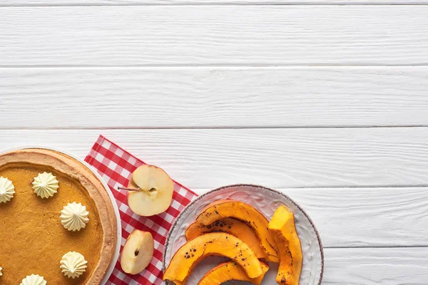 Deliziosa torta di zucca con panna montata su tovagliolo a quadretti vicino alla zucca cotta a fette e mela tagliata su tavolo di legno bianco — Foto stock