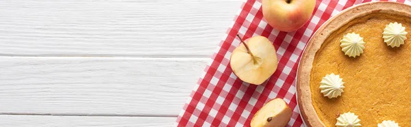 Torta di zucca saporita con panna montata su tovagliolo a quadretti vicino a taglio e mele intere su tavolo di legno bianco — Foto stock