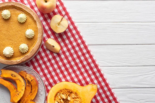 Tasty pumpkin pie with whipped cream on checkered napkin near raw and sliced baked pumpkins, cut and whole apples on white wooden table — Stock Photo