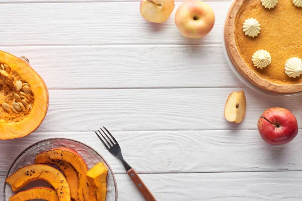 Tarte à la citrouille savoureuse avec crème fouettée près de la citrouille crue et tranchée cuite au four, pommes coupées et entières, et fourchette sur table en bois blanc — Photo de stock