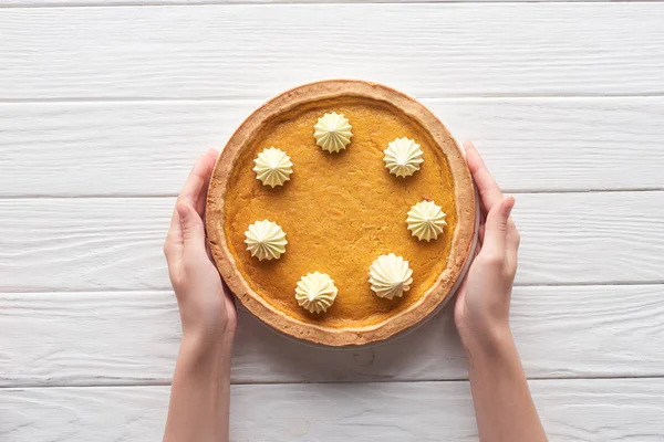 Vista recortada de las manos femeninas cerca de sabroso pastel de calabaza con crema batida en la mesa de madera blanca - foto de stock