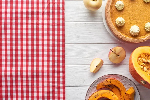 Leckere Kürbiskuchen mit Schlagsahne neben karierter Tischdecke, rohe und gebackene Kürbisse, ganze und geschnittene Äpfel auf weißem Holztisch — Stockfoto
