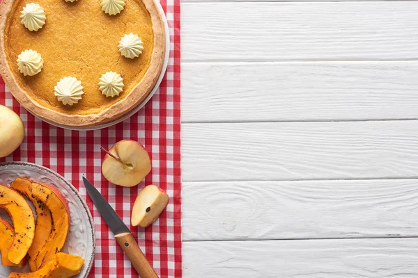 Leckere Kürbiskuchen mit Schlagsahne auf karierter Tischdecke neben gebackenem Kürbis, geschnittenen und ganzen Äpfeln und Messer auf weißem Holztisch — Stockfoto