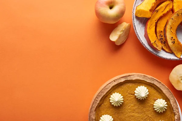 Deliziosa torta di zucca con panna montata vicino alla zucca al forno tagliata a fette, mele intere e tagliate sulla superficie arancione — Foto stock