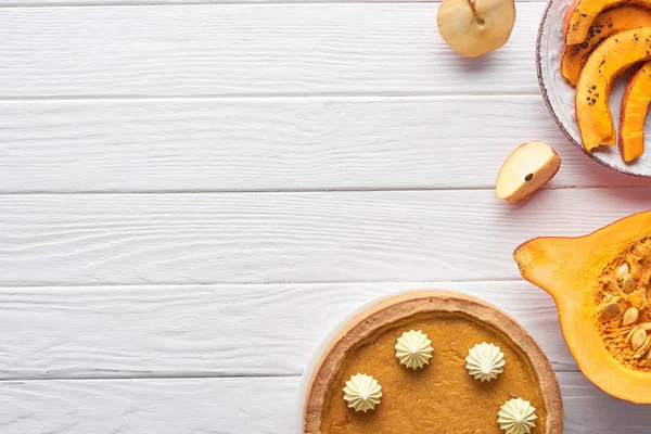 Вкусный тыквенный пирог со взбитыми сливками рядом с сырыми и запечёнными тыквами, целыми и разрезанными яблоками на белом деревянном столе — стоковое фото