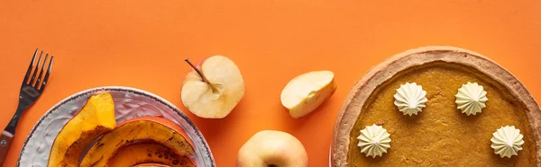 Plan panoramique de délicieuse tarte à la citrouille avec crème fouettée près de tranches de citrouille cuite au four, pommes entières et coupées, et fourchette sur la surface orange — Photo de stock