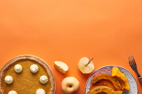 Délicieuse tarte à la citrouille avec crème fouettée près de tranches de citrouille cuite au four, pommes entières et coupées, et fourchette sur la surface orange — Photo de stock