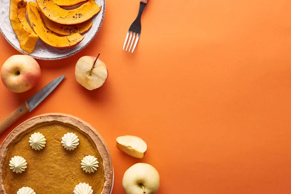 Вкусный тыквенный пирог со взбитыми сливками рядом с нарезанной запечённой тыквой, целые и нарезанные яблоки, нож и вилка на оранжевой поверхности — стоковое фото