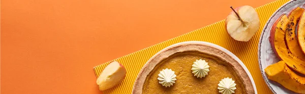 Plan panoramique de délicieuse tarte à la citrouille avec crème fouettée près tranchée citrouille cuite au four et pomme coupée sur la surface orange — Photo de stock