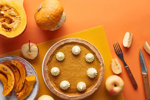 Вкусный тыквенный пирог со взбитыми сливками рядом с сырыми и запечёнными тыквами, целыми и разрезанными яблоками, ножом и вилкой на оранжевой поверхности — стоковое фото