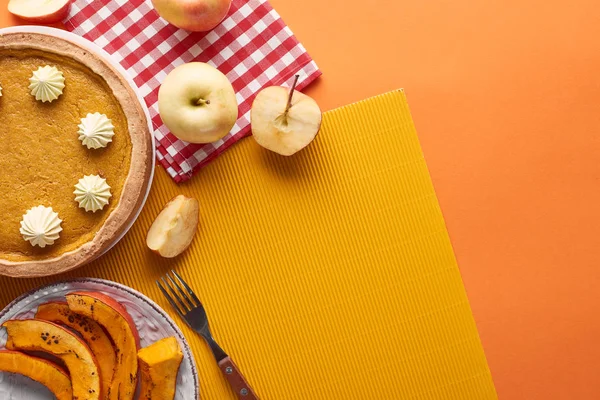 Вкусный тыквенный пирог со взбитыми сливками рядом с нарезанной печеной тыквой, целые и нарезанные яблоки, салфетки, нож и вилка на оранжевой поверхности — стоковое фото