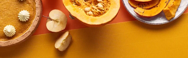 Panoramic shot of pumpkin pie with whipped cream near raw and baked pumpkins, cut apples, and textured napkin on orange surface — Stock Photo