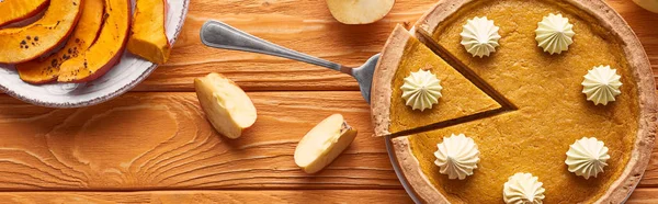Plano panorámico de delicioso pastel de calabaza con crema batida cerca de calabaza horneada en rodajas y manzana cortada en una mesa de madera naranja - foto de stock