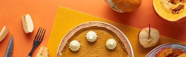 Panoramic shot of delicious pumpkin pie with whipped cream near raw and baked pumpkins, cut apple, knife and fork on orange surface — Stock Photo