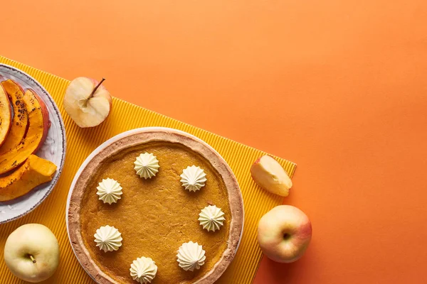 Delicious pumpkin pie with whipped cream on textured napkin near sliced baked pumpkin, cut and whole apples on orange surface — Stock Photo