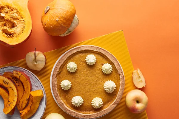 Вкусный тыквенный пирог со взбитыми сливками на текстурной салфетке рядом с сырыми и запечёнными тыквами, вырезанными и целыми яблоками на оранжевой поверхности — стоковое фото