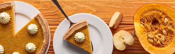 Plano panorámico de delicioso pastel de calabaza con crema batida cerca de la mitad de la calabaza cruda, y manzana cortada en una mesa de madera naranja - foto de stock