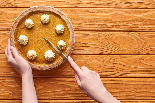 Vista ritagliata della donna che taglia deliziosa torta di zucca con panna montata sul tavolo di legno arancione — Foto stock