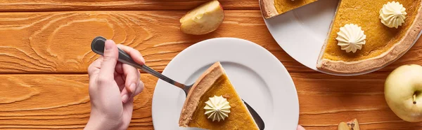 Cropped view of woman holding spatula with piece of pumpkin pie with whipped cream near cut and whole apples on orange wooden table, panoramic shot — Stock Photo