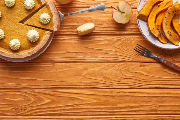 Delicioso pastel de calabaza con crema batida cerca de calabaza horneada en rodajas, manzana cortada y tenedor en la mesa de madera naranja - foto de stock