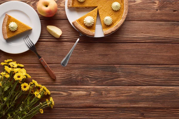 Delicioso pastel de calabaza con crema batida en platos con espátula y tenedor cerca de manzanas cortadas y enteras, y flores amarillas en mesa de madera marrón - foto de stock