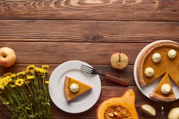 Вкусный тыквенный пирог со взбитыми сливками рядом с вилкой, половина сырой тыквы, срезанные и целые яблоки, и желтые цветы на коричневом деревянном столе — стоковое фото