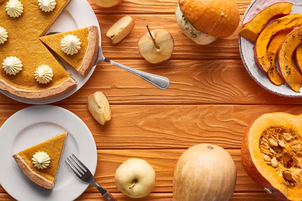 Délicieuse tarte à la citrouille avec crème fouettée sur assiettes avec spatule et fourchette près de pommes coupées et entières, citrouilles cuites et crues sur table en bois orange — Photo de stock