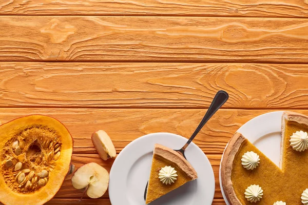 Delicioso pastel de calabaza con crema batida cerca de cortar y manzanas enteras, la mitad de la calabaza cruda en la mesa de madera naranja - foto de stock