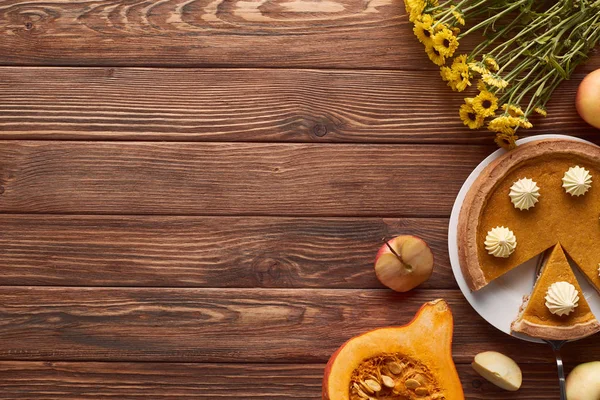 Sabroso pastel de calabaza con crema batida cerca de manzanas enteras y cortadas, la mitad de la calabaza cruda y flores amarillas en la superficie de madera marrón - foto de stock