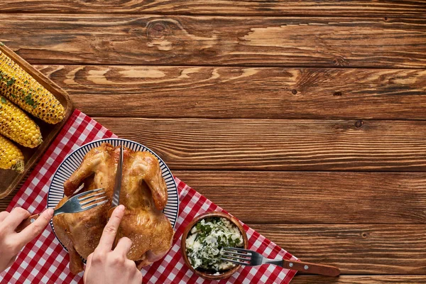 Cropped view of woman cutting roasted turkey on red plaid napkin near grilled corn on wooden table — Stock Photo