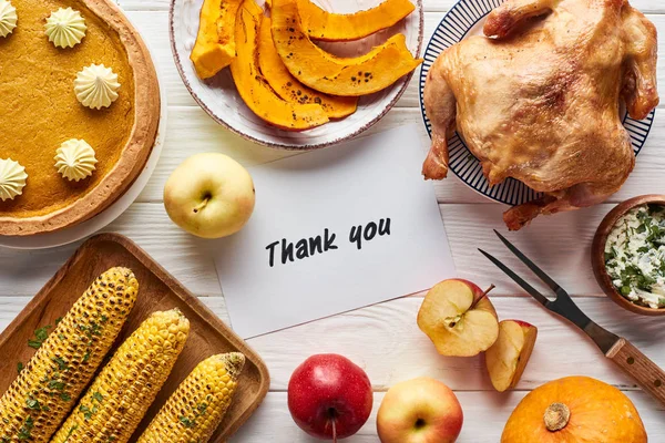 Vue du dessus de dinde rôtie, tarte à la citrouille et légumes grillés servis sur une table en bois blanc près de la carte de remerciement — Photo de stock