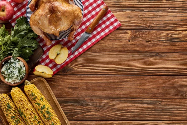 Vista superior del pavo asado, maíz a la parrilla, manzanas, perejil y flores silvestres amarillas en la servilleta a cuadros roja en la mesa de madera - foto de stock