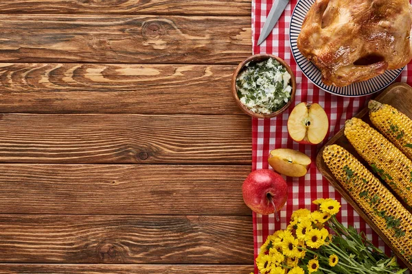 Vista superior del pavo asado, maíz a la parrilla, manzanas y flores silvestres amarillas en la servilleta a cuadros roja en la mesa de madera - foto de stock