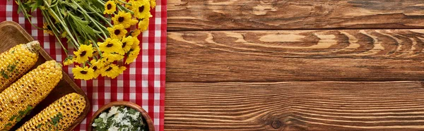 Top view of grilled corn and yellow wildflowers on red checkered napkin on wooden table, panoramic shot — Stock Photo