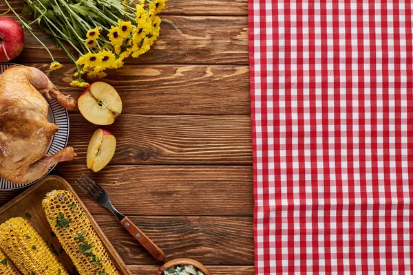 Верхний вид жареной индейки, жареной кукурузы, яблок и желтых полевых цветов возле красной клетчатой салфетки на деревянном столе — стоковое фото