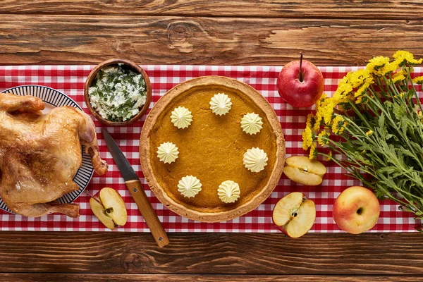Top view of roasted turkey, pumpkin pie and yellow flowers served on wooden table with red plaid napkin — Stock Photo