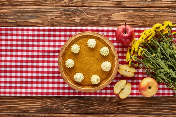 Vista superior del pastel de calabaza, manzanas y flores amarillas servidas en mesa de madera con servilleta a cuadros roja - foto de stock