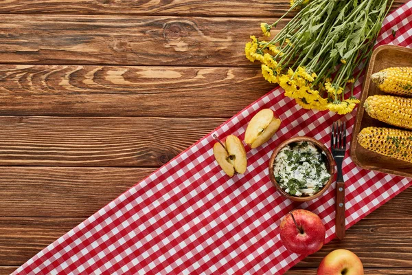 Vue de dessus de maïs grillé, pommes et fleurs jaunes servis sur table en bois sur serviette rouge — Photo de stock