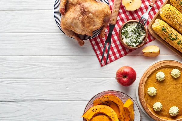 Vista superior de pavo asado, pastel de calabaza y maíz a la parrilla servido en mesa de madera blanca con servilleta a cuadros roja - foto de stock