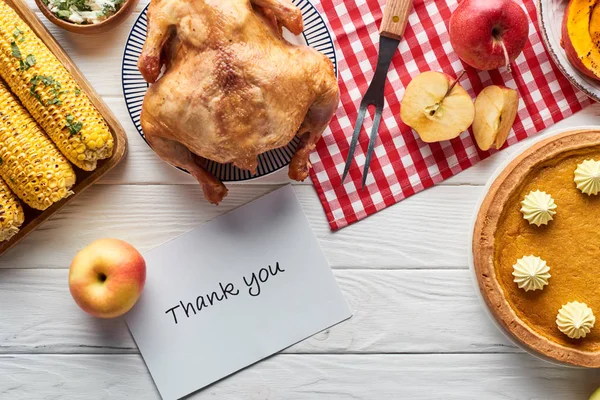 Top view of roasted turkey, pumpkin pie and grilled vegetables served on white wooden table with red plaid napkin and thank you card — Stock Photo