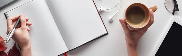 Visão parcial da mulher segurando xícara de café e caneta perto de notebook, fones de ouvido, tablet digital, óculos e folhas vermelhas na mesa branca, tiro panorâmico — Fotografia de Stock