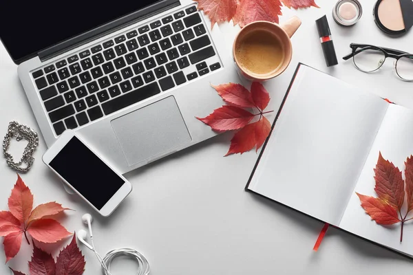 Vista superior de la computadora portátil cerca de teléfono inteligente, taza de café, cosméticos, auriculares, vasos, portátil y hojas rojas de uvas silvestres en la mesa blanca - foto de stock