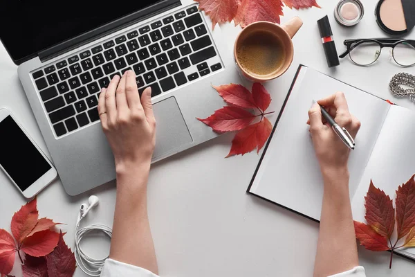 Cropped view of woman using laptop and holding pen near notebook, cosmetics, glasses, earphones, smartphone and red leaves of wild grapes on white table — Stock Photo