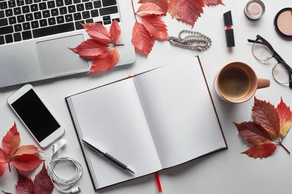 Vista superior de laptop perto de smartphone, xícara de café, cosméticos, fones de ouvido, óculos, notebook e folhas vermelhas de uvas selvagens na mesa branca — Fotografia de Stock