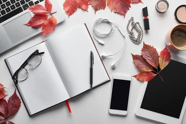 Vista superior de la computadora portátil cerca de teléfono inteligente, tableta digital, taza de café, cosméticos, auriculares, vasos, portátil y hojas rojas de uvas silvestres en la mesa blanca - foto de stock