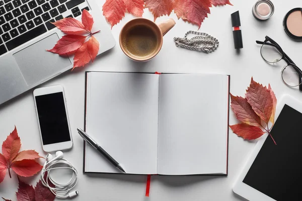 Vista dall'alto del computer portatile vicino smartphone, tablet digitale, tazza di caffè, cosmetici, auricolari, occhiali, notebook e foglie rosse sul tavolo bianco — Foto stock