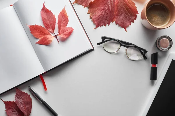 Верхний вид ноутбука возле стаканов, чашки кофе, косметики, ручки и красные листья дикого винограда на белом столе — стоковое фото