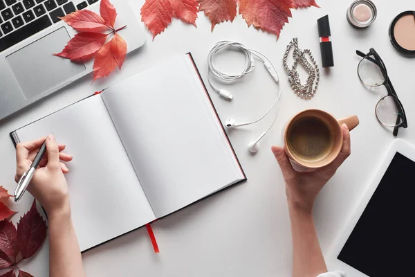 Vista ritagliata di donna in possesso di tazza di caffè e penna vicino notebook, cosmetici, auricolari, occhiali, tablet digitale e foglie rosse sul tavolo bianco — Foto stock